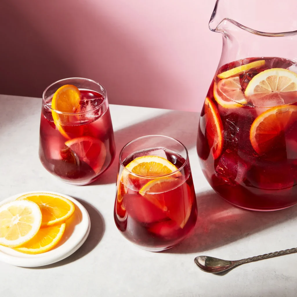 "Refreshing red sangria with slices of oranges and lemons in a glass pitcher and two glasses against a pink background"