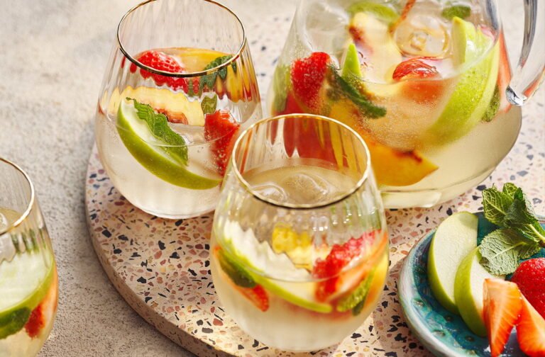 Refreshing white sangria with slices of green apple, strawberries, and mint leaves in a glass pitcher and three glasses on a terrazzo serving board