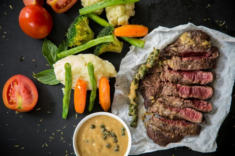 Grilled beef steak slices with creamy peppercorn sauce, mashed potatoes, and a medley of vegetables including broccoli, carrots, green beans, and tomatoes on a dark background