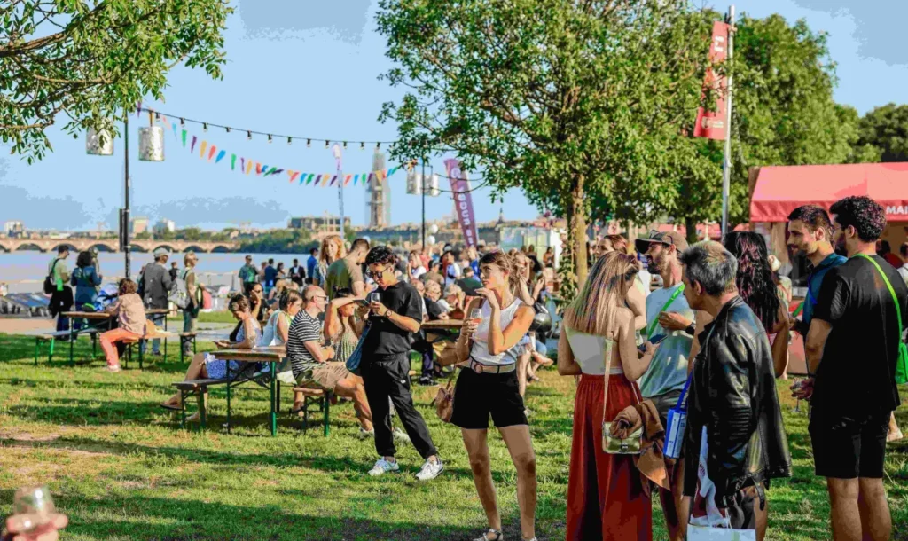 Crowd enjoying the Bordeaux Wine Festival by the waterfront with colorful decorations and scenic views. Attendees are tasting wine and socializing in a vibrant outdoor setting.