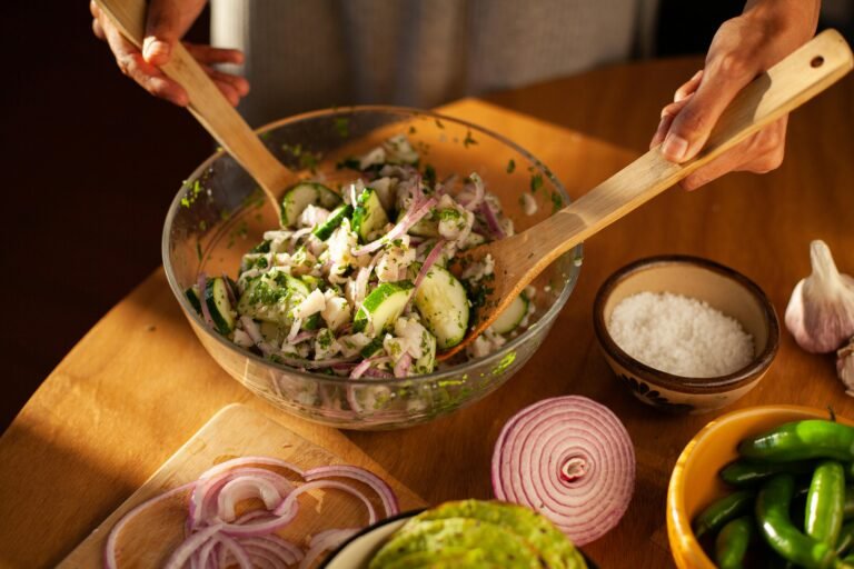 Person preparing a fresh cucumber and red onion salad with cilantro in a glass bowl, surrounded by sliced red onion, green peppers, and garlic on a wooden table