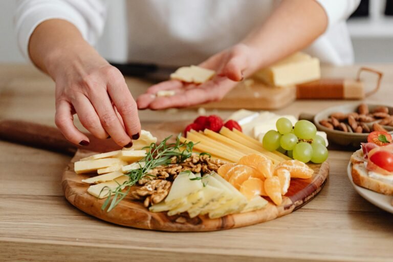 Person preparing a cheese platter with a variety of cheeses, fresh fruits, nuts, and herbs on a wooden board