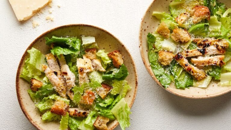 Two plates of classic Caesar salad featuring crisp romaine lettuce, grilled chicken strips, crunchy croutons, and a generous sprinkle of grated Parmesan cheese, arranged on a light-colored table.