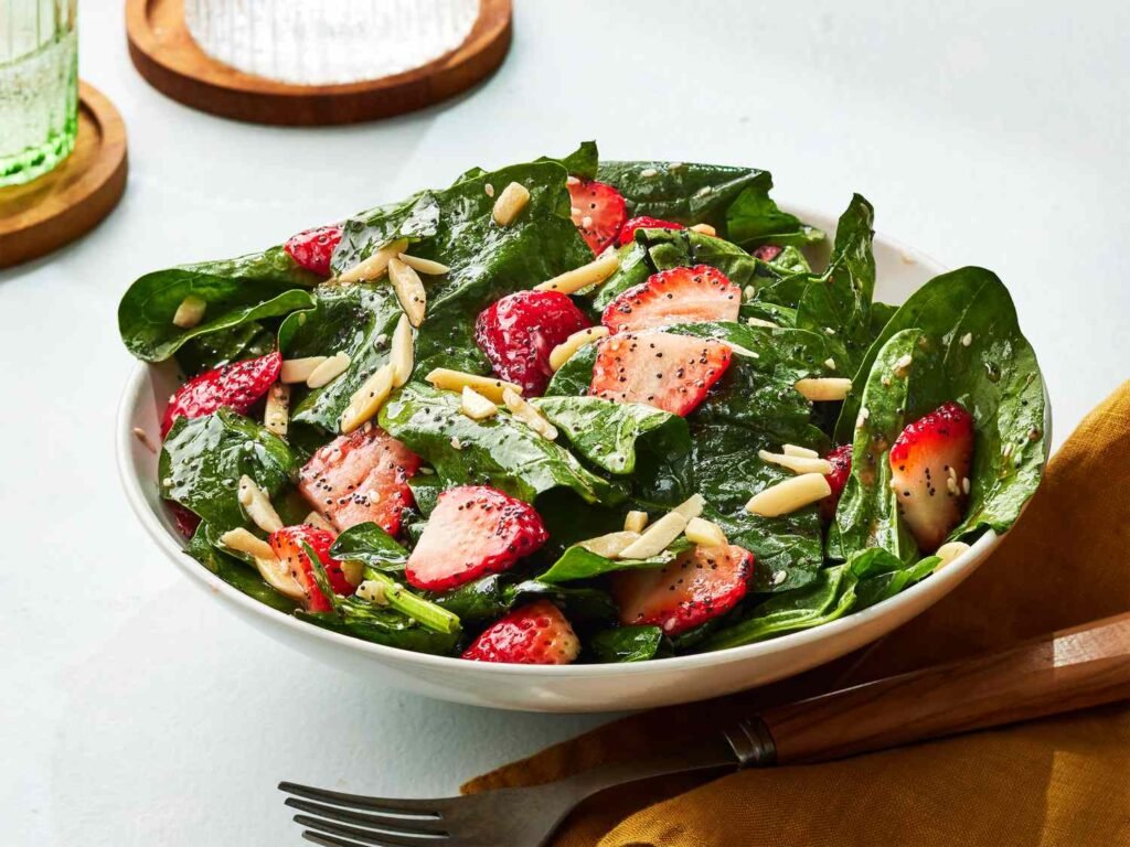 A fresh strawberry spinach salad in a white bowl, adorned with sliced strawberries, fresh spinach leaves, slivered almonds, and drizzled with a poppy seed dressing, set on a light-colored surface with a fork and a yellow napkin.