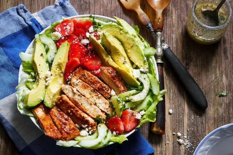 A nutritious grilled chicken salad served in a white bowl, featuring slices of grilled chicken breast, fresh avocado, cucumbers, tomatoes, and crumbled feta cheese, placed on a rustic wooden table with a blue checkered napkin.