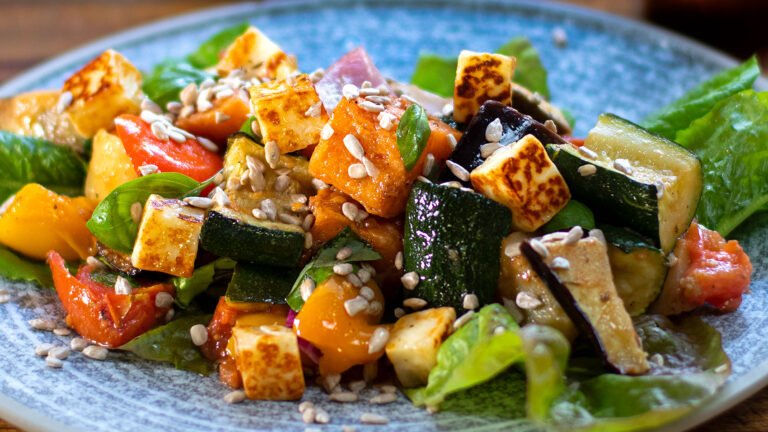 A close-up view of a colorful roasted vegetable salad featuring grilled zucchini, bell peppers, tomatoes, and cubes of tofu, garnished with sunflower seeds and fresh basil leaves on a blue speckled plate.