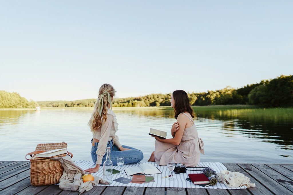 Two women having a picnic by the lake with books and a wicker basket.
