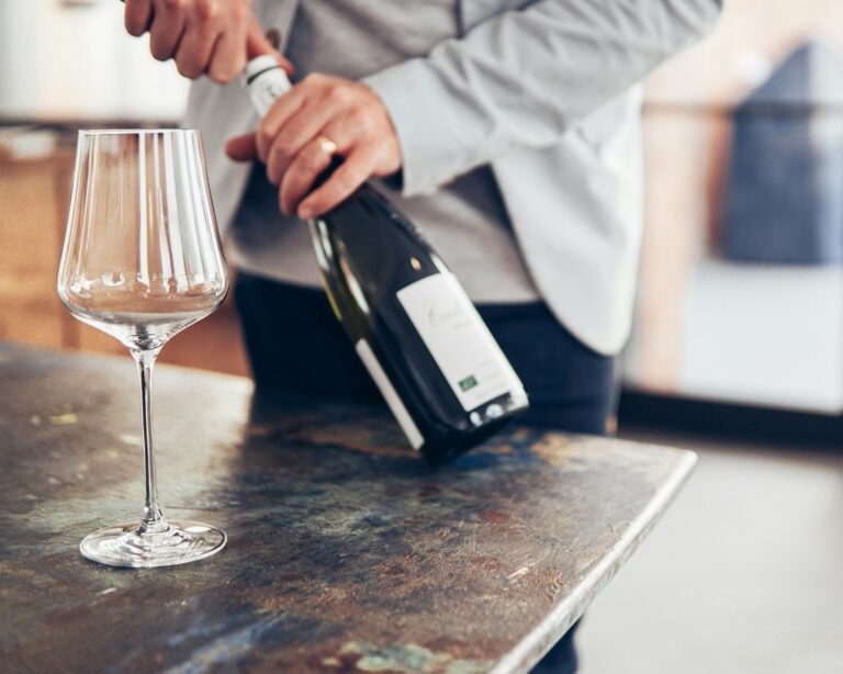 Person opening a bottle of wine with an empty wine glass on the table.