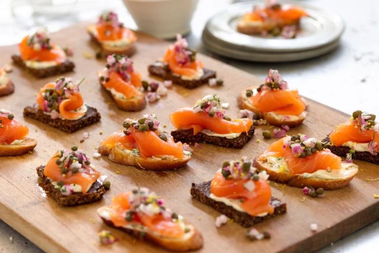 A wooden platter with smoked salmon canapés topped with capers and herbs, perfect for a summer appetizer.