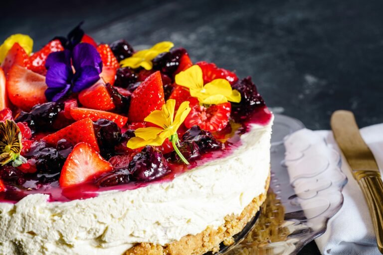 Cheesecake topped with fresh strawberries, blackberries, and edible flowers on a dark background.