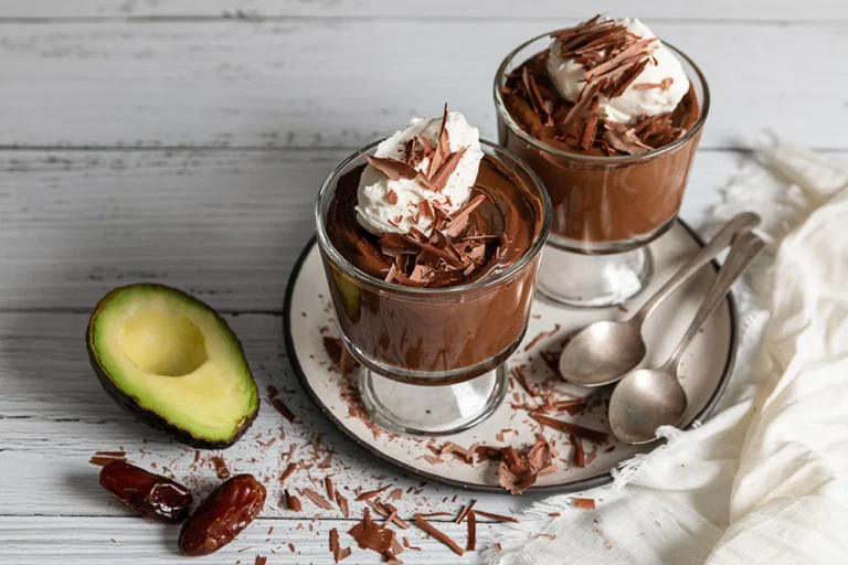 Two glasses of creamy chocolate mousse topped with whipped cream and chocolate shavings, with an avocado and dates on the side.