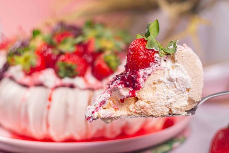 Close-up of a slice of pavlova topped with fresh strawberries and berry compote, with the rest of the pavlova in the background.