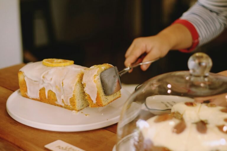 Lemon polenta cake being sliced, topped with a lemon slice, served on a white plate with a cake cover in the foreground.