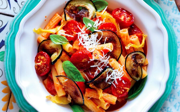 Colorful plate of pasta alla norma with fresh basil, eggplant, cherry tomatoes, and grated cheese.