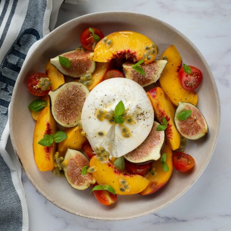 Vibrant summer salad featuring fresh figs, peach slices, cherry tomatoes, and burrata cheese garnished with basil leaves, served in a ceramic bowl.