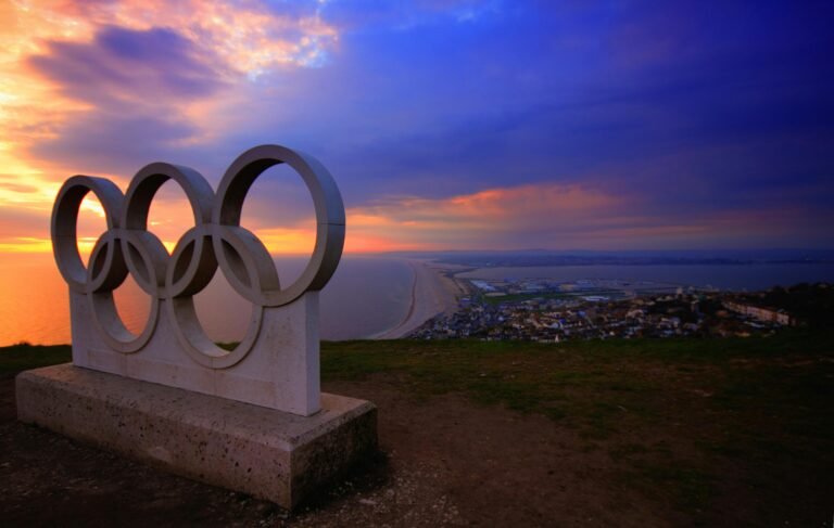 Olympic rings sculpture overlooking a coastal city at sunset.