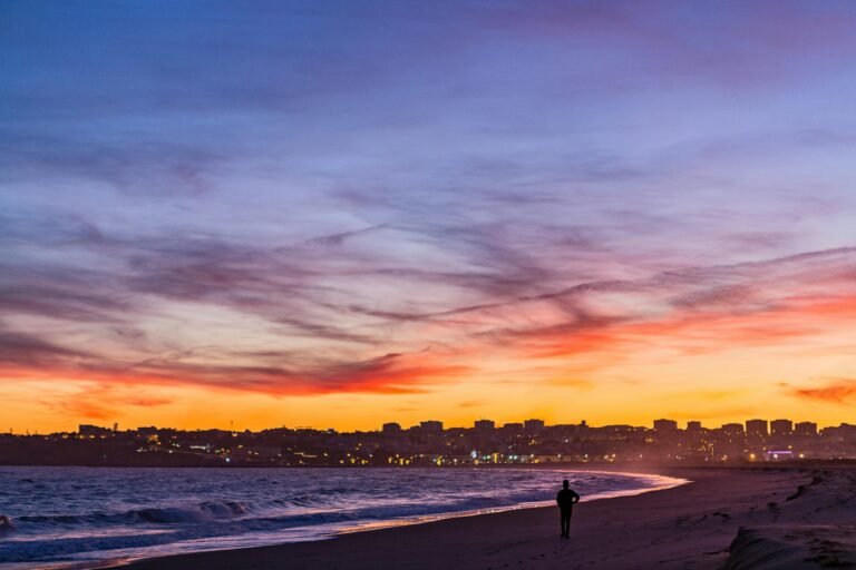 A solitary figure walking along a beach at sunset with a city skyline in the distance.