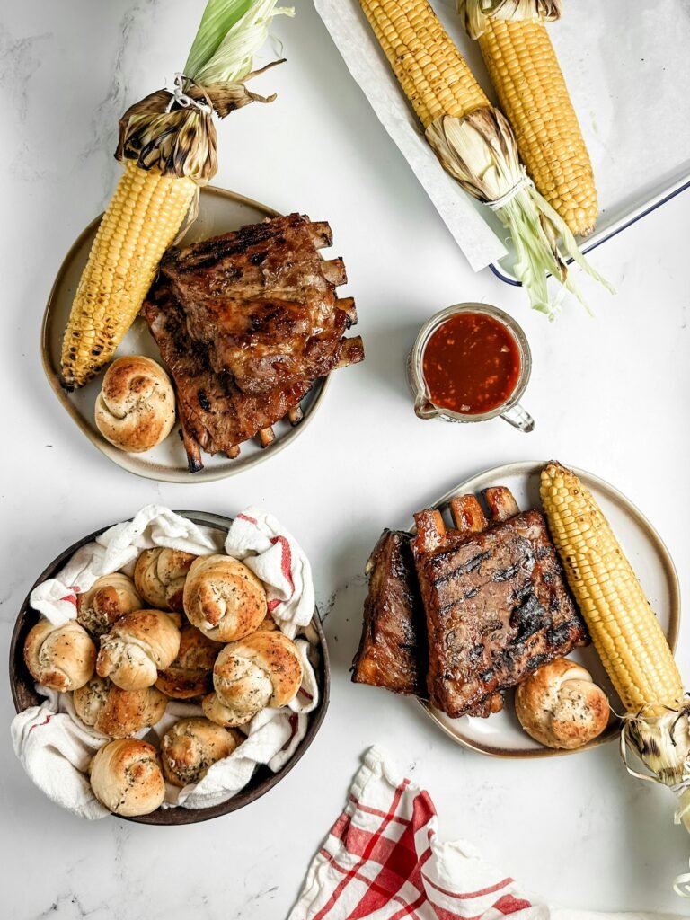 Mouthwatering plate of barbecue pork ribs served with a corn cob, barbecue sauce, and a freshly baked bread roll.