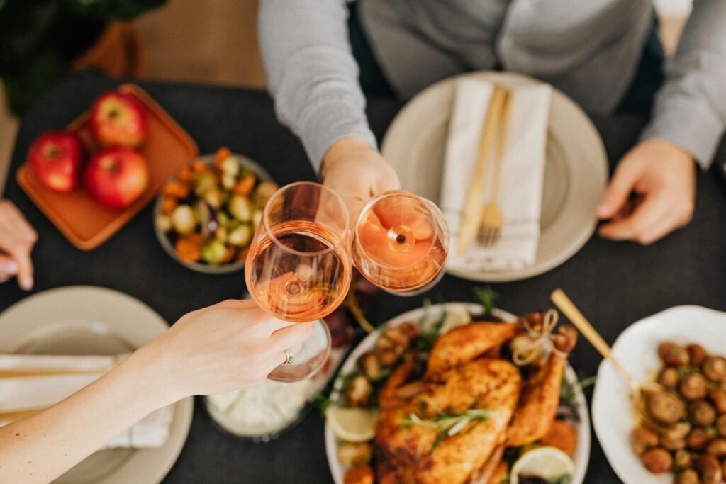 Two people toasting with glasses of rosé wine over a dinner table filled with roasted chicken and vegetables.