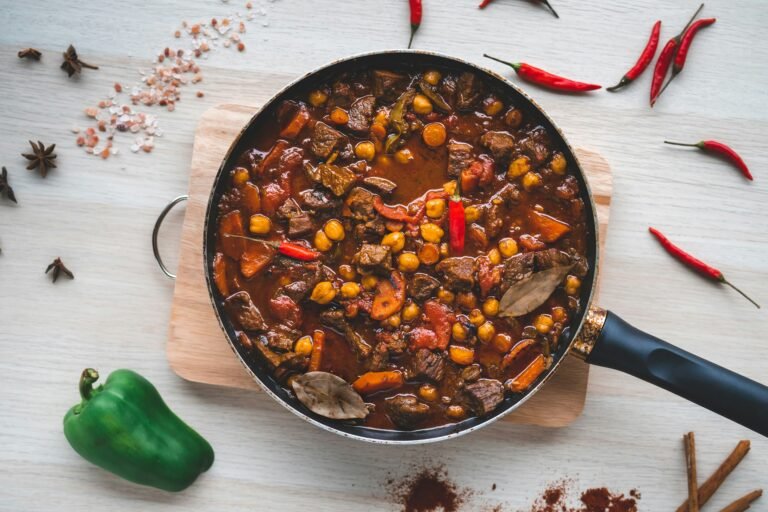 A pan of rich beef stew with chickpeas and vegetables, surrounded by chili peppers and spices.