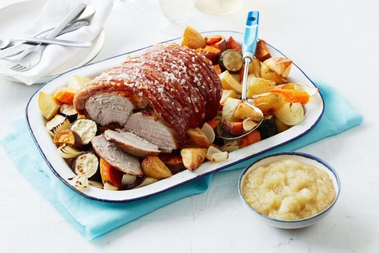 Roast pork with crispy crackling served on a platter with roasted vegetables and a side of applesauce