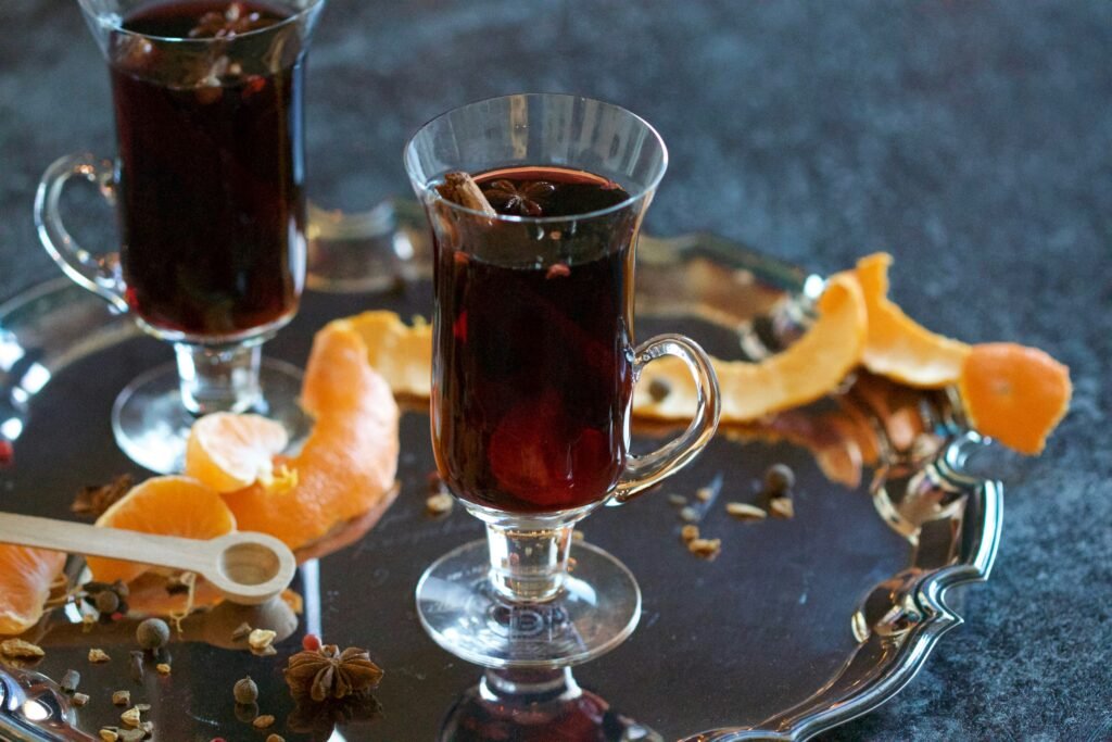 Glass cups of spiced mulled wine with cinnamon sticks, surrounded by orange peel and star anise on a silver tray.