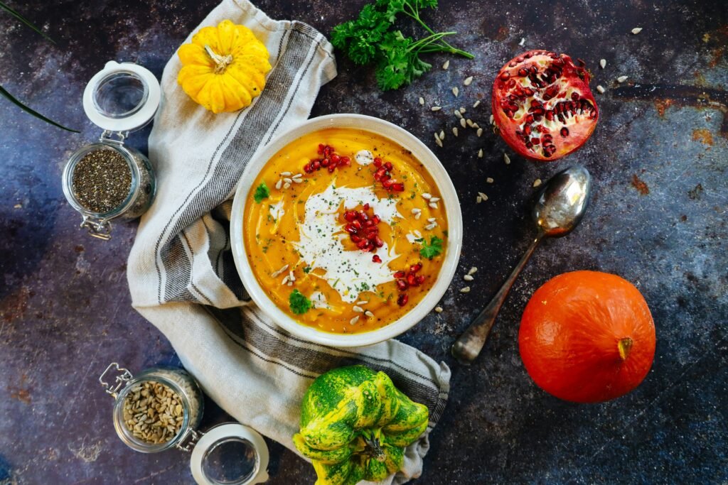 A bowl of creamy pumpkin soup garnished with pomegranate seeds and herbs, surrounded by autumnal vegetables and seeds.