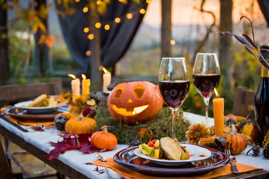 Halloween-themed dinner table with a carved pumpkin, candles, and glasses of red wine, set against a twilight backdrop.