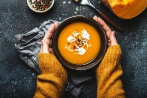 Hands holding a bowl of creamy pumpkin soup with seeds and spices