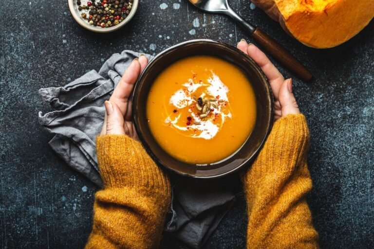 Hands holding a bowl of creamy pumpkin soup with seeds and spices