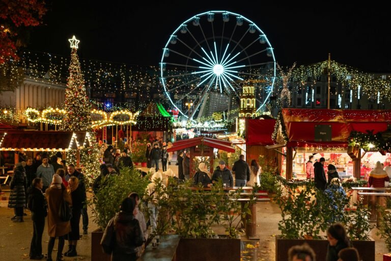 Christmas market at night with festive lights, decorated trees, and a Ferris wheel, bustling with visitors enjoying holiday festivities.