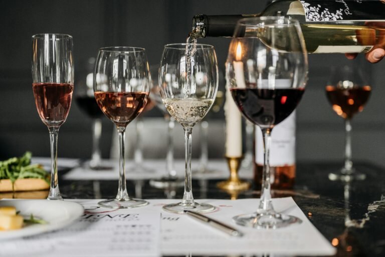 Close-up of a hand pouring white wine into a glass among other filled wine glasses, with a tasting notes sheet and cheese in the background.