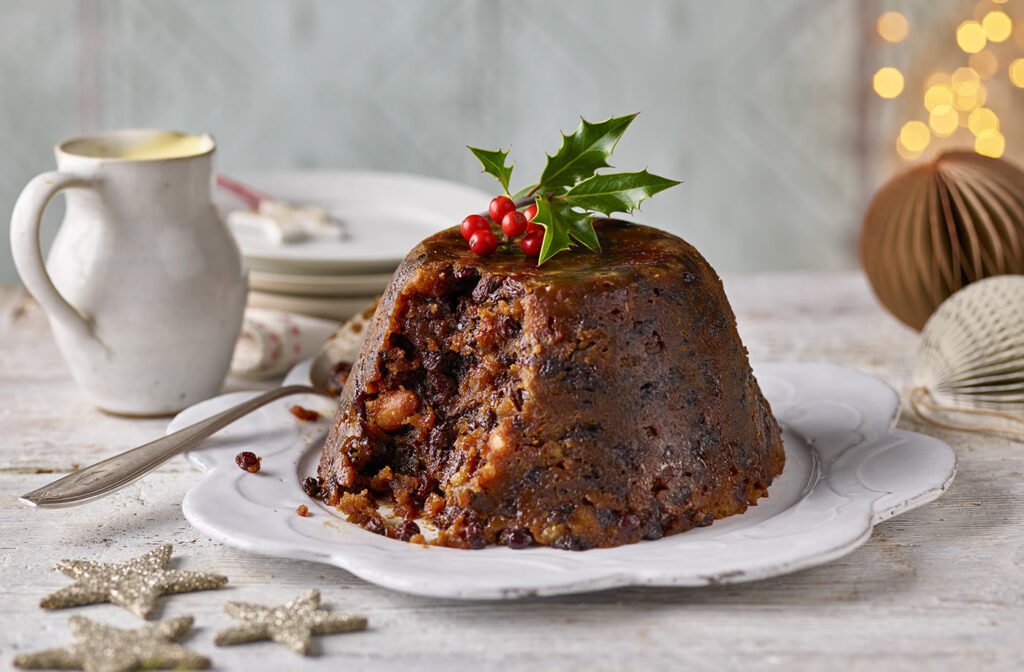 Traditional Christmas pudding garnished with holly on a white plate, served with a jug of cream, set on a festive table.