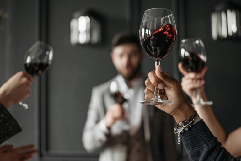 A group of people raising red wine glasses in a celebratory toast, with a blurred man in a suit in the background, creating a sophisticated atmosphere.