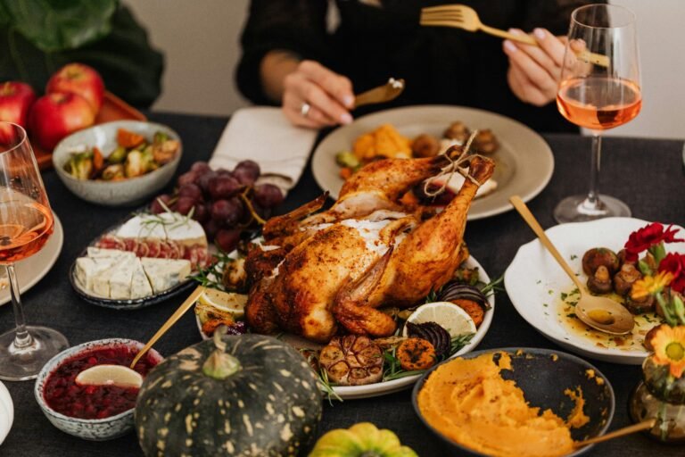 A beautifully roasted turkey surrounded by classic Thanksgiving side dishes, including mashed sweet potatoes, cheese platter, fresh grapes, cranberry sauce, and roasted vegetables, with glasses of wine on a dining table.