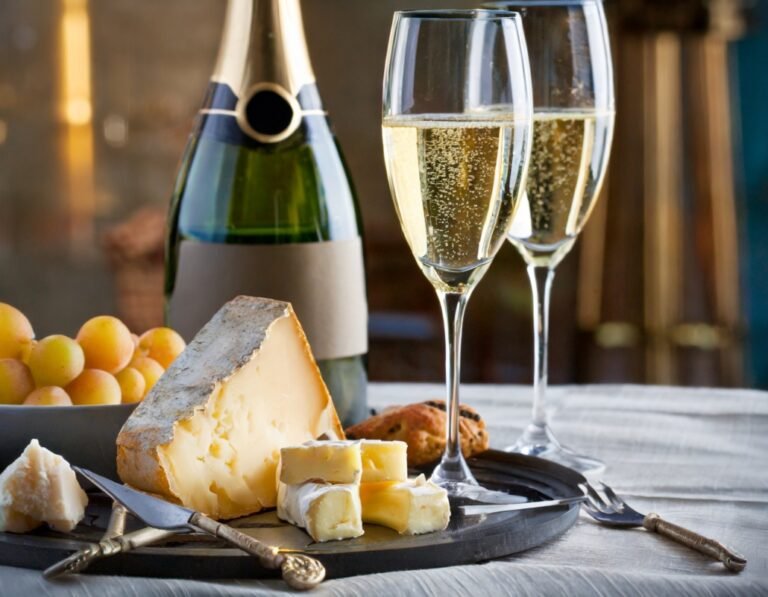 Champagne and cheese setup featuring a bottle of champagne, two glasses of bubbly, assorted cheeses, and a bunch of grapes on a tray, in a cozy, indoor setting.