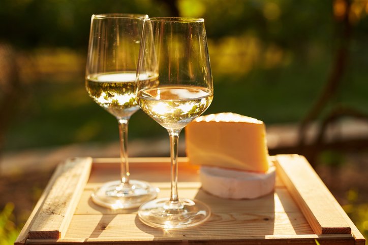 Close-up of two glasses of chardonnay and a selection of cheeses on a wooden tray, basking in warm sunlight in an outdoor garden setting.