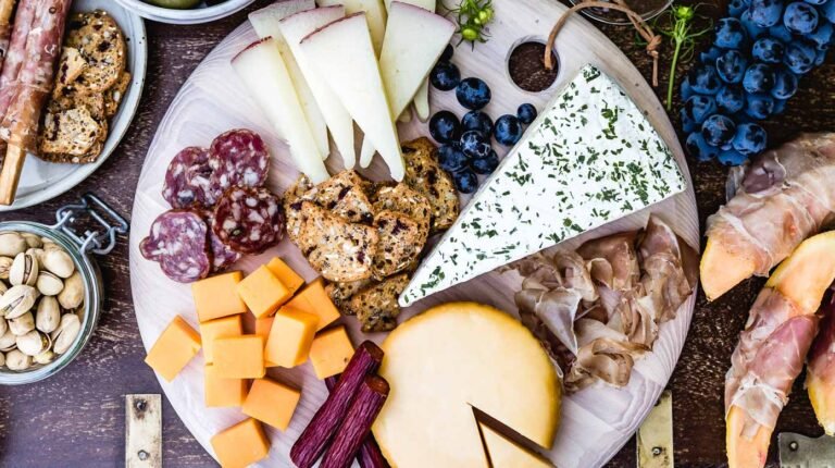 A charcuterie board featuring a variety of cheeses, crackers, cured meats, fresh blueberries, and pistachios arranged elegantly on a wooden board, perfect for a gathering or wine pairing.