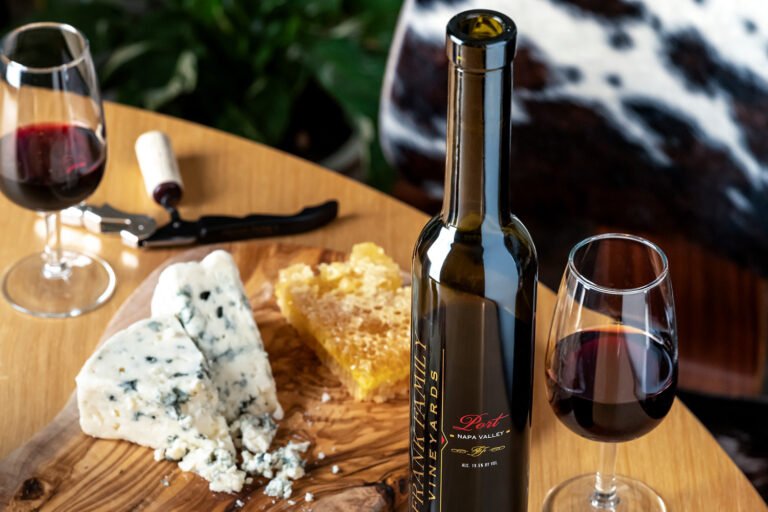 A bottle of Frank Family Vineyards Napa Valley Port with two glasses of red wine, accompanied by a wedge of blue cheese and a piece of honeycomb on a wooden board.