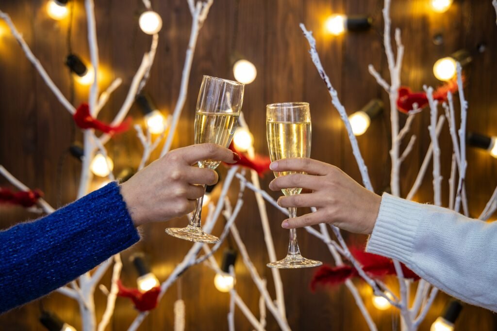 Two hands clinking champagne glasses in a festive setting with decorative white branches, red ribbons, and warm string lights in the background.