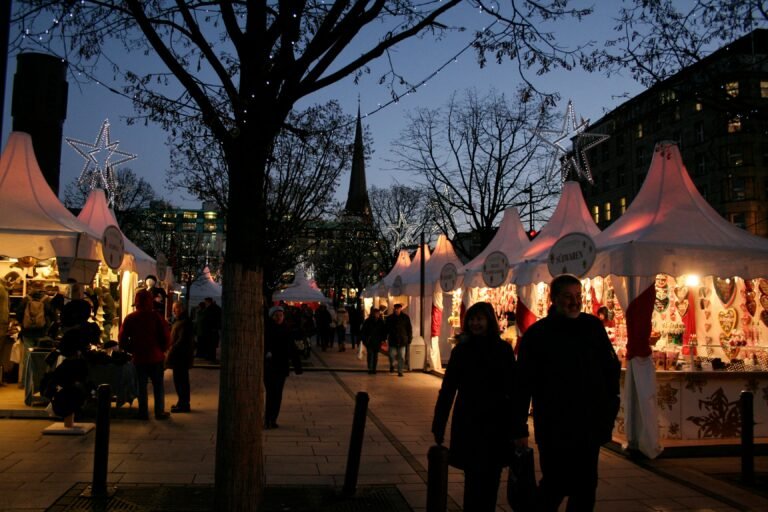 Illuminated Christmas market tents at dusk, with people strolling along the festive stalls decorated with holiday lights and stars.