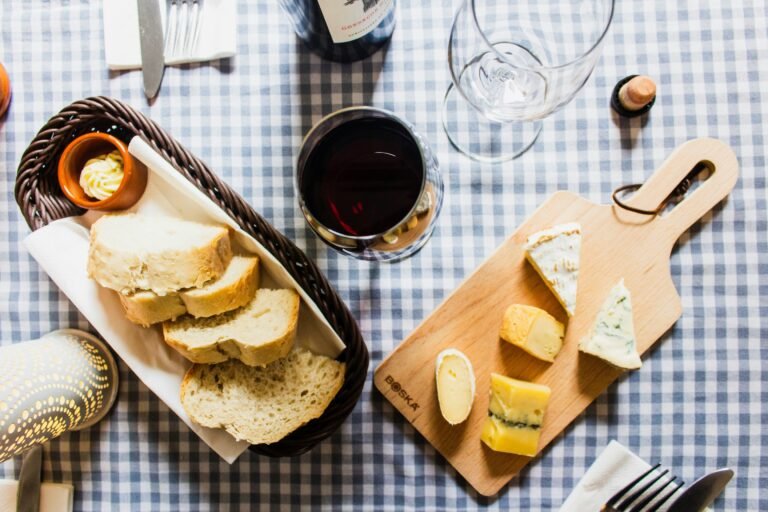 Overhead view of a rustic cheese and wine setup featuring sliced bread in a basket, assorted cheeses on a wooden board, and a glass of red wine on a checkered tablecloth.