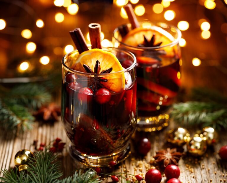 Two glasses of festive mulled wine with orange slices, cinnamon sticks, cranberries, and star anise, surrounded by holiday decorations and twinkling lights.