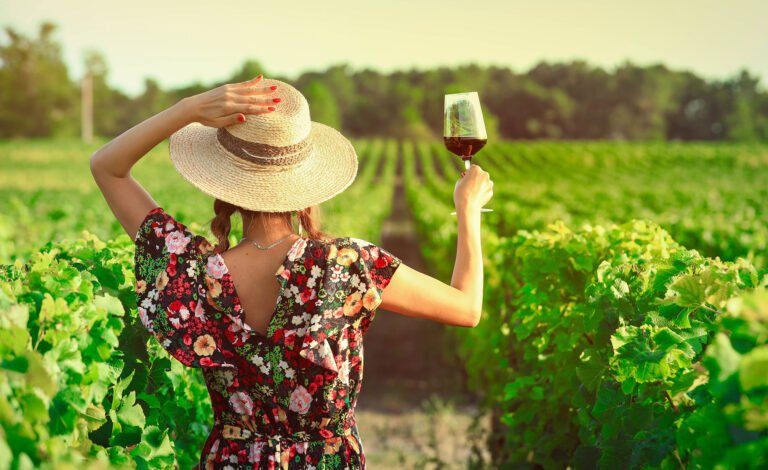 Woman in a floral dress holding a glass of red wine while enjoying a sunny vineyard view.