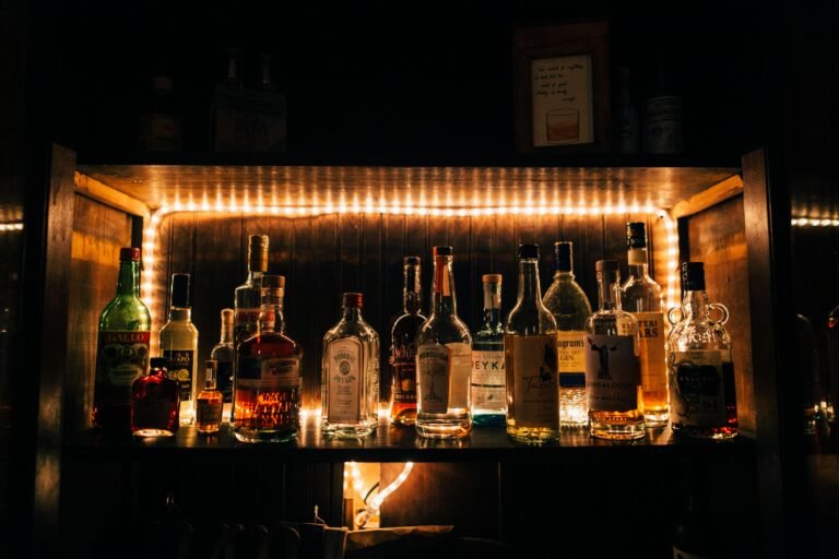 A dimly lit bar shelf displaying an assortment of liquor bottles illuminated by warm string lights.