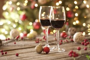 Two glasses of red wine on a rustic wooden table with festive Christmas decorations and lights in the background.