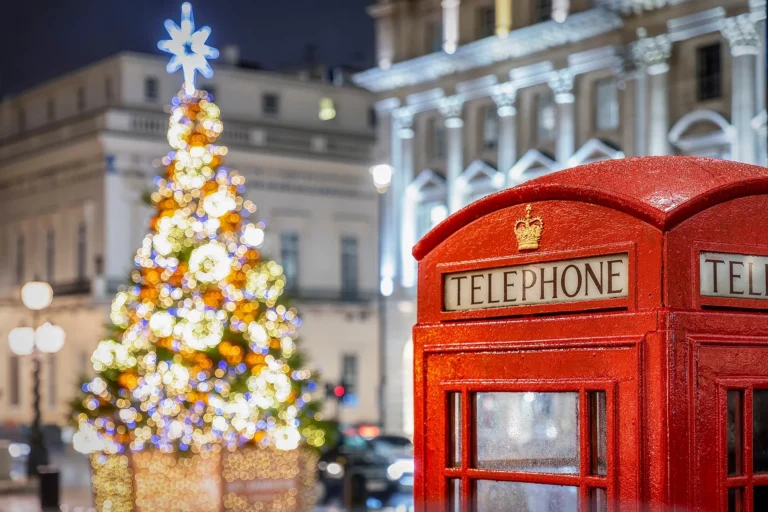 A classic red British telephone booth with a festive Christmas tree and glowing lights in the background at night.