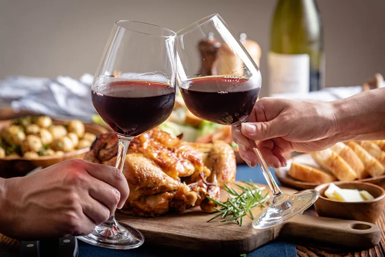 Two people clinking glasses of red wine at a dinner table with roasted chicken and side dishes.