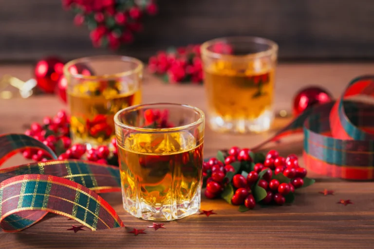 Three glasses of whiskey on a festive wooden table decorated with red berries, plaid ribbons, and holiday ornaments.