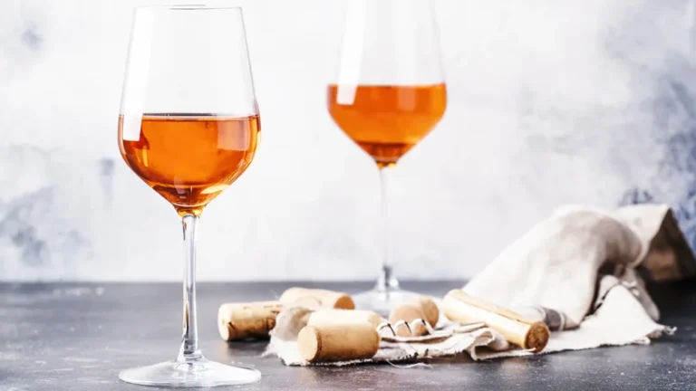 Two glasses of orange wine on a grey surface with wine corks and a linen napkin, highlighting the rich amber hue of the drink.